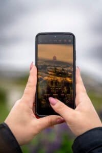 Hands with painted nails holding smartphone capturing a rocky hill with a red and white lighthouse.