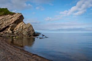 Rocky coastline with large rock formation, calm water, sandy shore, and partly cloudy sky.