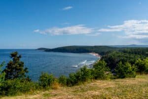 Coastal view with blue sea, sandy beach, green shrubs, forested hills, and a partly cloudy sky.