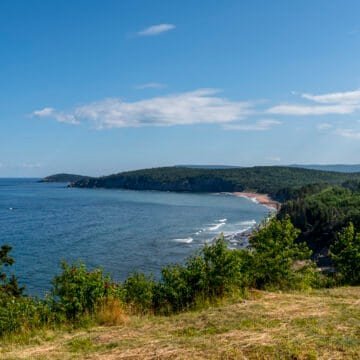 Coastal view with blue sea, sandy beach, green shrubs, forested hills, and a partly cloudy sky.