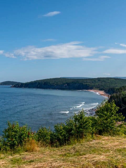 Coastal view with blue sea, sandy beach, green shrubs, forested hills, and a partly cloudy sky.