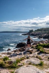 Woman stands on rock by rocky coastline with ocean, waves, forest, and partly cloudy sky.
