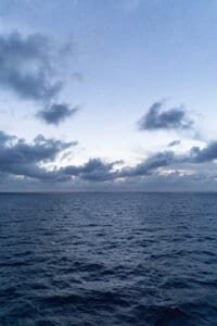 Calm ocean under a blue sky with scattered dark clouds and a crescent moon on the horizon