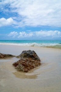 Sandy beach with rocks in shallow water, waves, turquoise ocean, and partly cloudy sky.