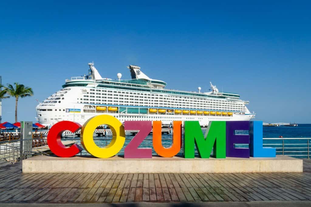 An image of a cruise ship with a sign that says Cozumel in the foreground with blue sky and a blue ocean