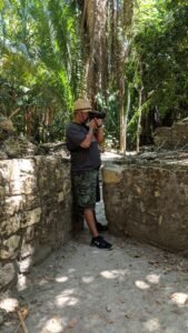 Person wearing straw hat and camouflage shorts taking photo near ancient stone walls in forest.