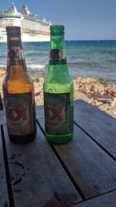 Two Dos Equis beer bottles on a wooden table by a rocky shore with ocean and cruise ship.