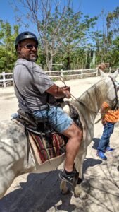 Man wearing a helmet and sunglasses riding a white horse with a decorative saddle blanket.