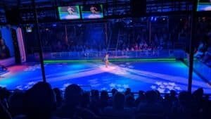 Ice skater performing on star-lit rink with audience and screens showing close-up views.