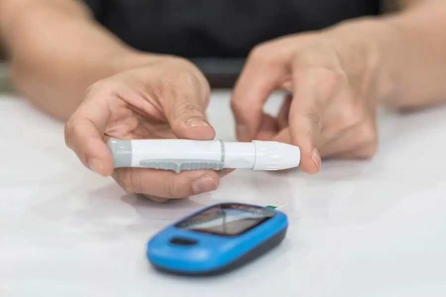A person prepares to use a lancet device for a blood glucose test, with a glucose meter placed on a white surface in front of them.