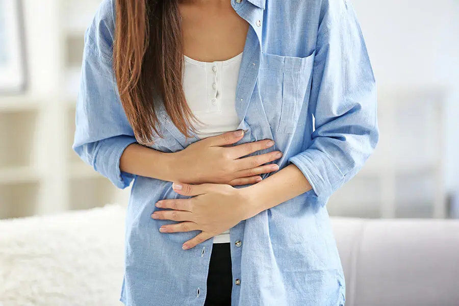 A woman wearing a blue shirt and white top is holding her stomach with both hands, suggesting abdominal discomfort or pain.