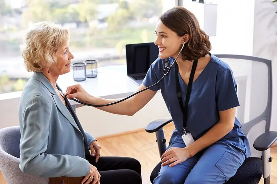 A healthcare professional in scrubs uses a stethoscope to listen to the chest of an older woman seated in an office.