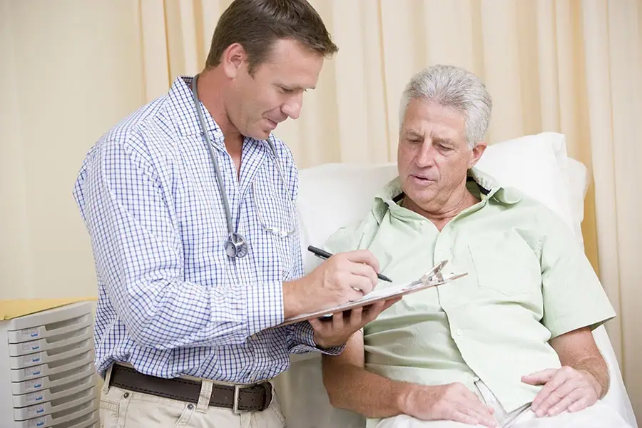 A doctor stands beside an older male patient sitting on an exam table, writing on a clipboard while they discuss medical information.