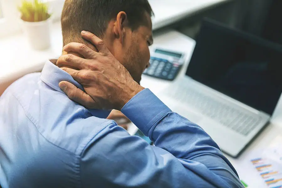 A man in a blue shirt sits at a desk with a laptop and paperwork, rubbing his neck as if experiencing discomfort or pain.