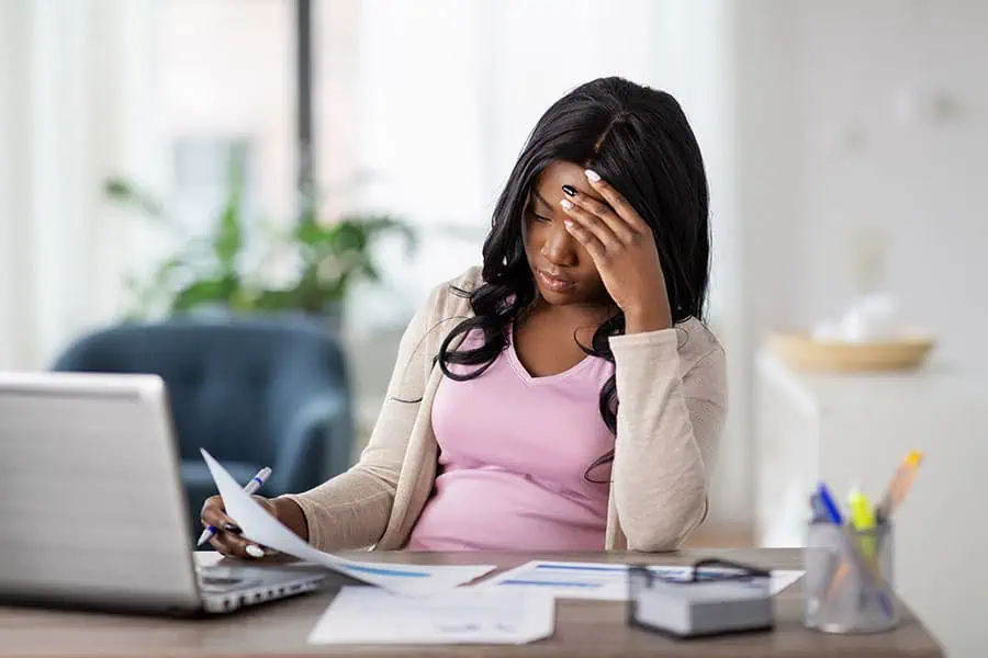 A woman sits at a desk with a laptop and papers, holding her head with one hand and looking stressed or tired.