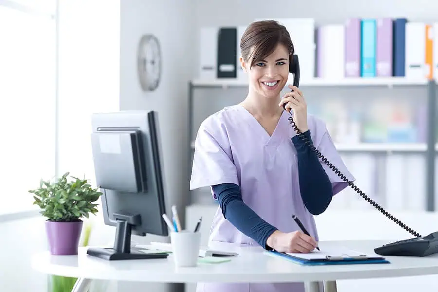 A nurse in scrubs stands at a desk, talking on a corded phone while writing on a clipboard, with a computer and potted plant nearby in a medical office.