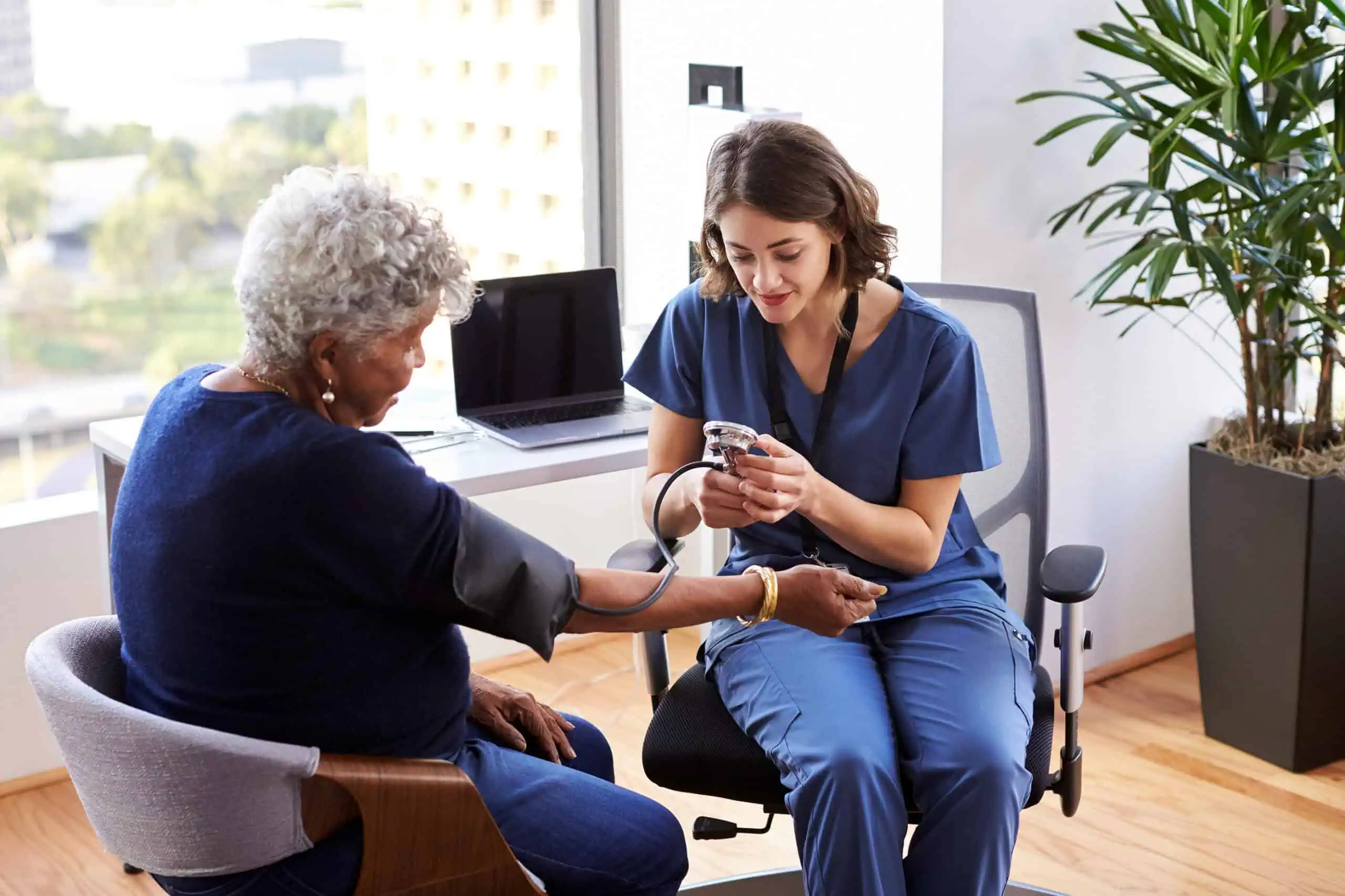 A nurse in scrubs measures the blood pressure of an older woman in an office setting with a laptop and plant visible in the background.
