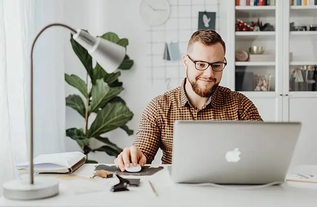 A man wearing glasses works at a desk with a laptop, books, and papers; a lamp and plant are visible in the background.