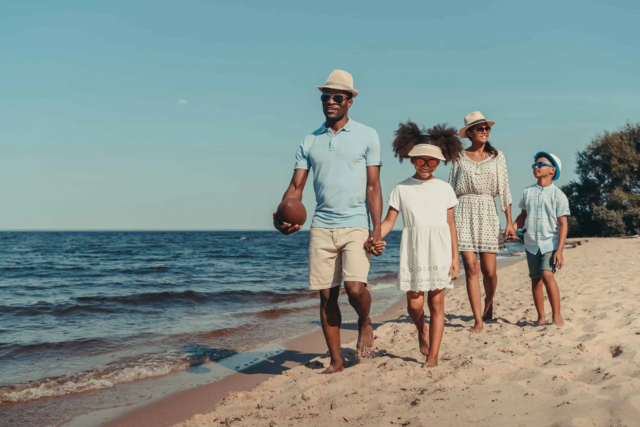 A family of four walks along a sandy beach near the water on a sunny day; the father holds a football and everyone wears summer clothing and hats.