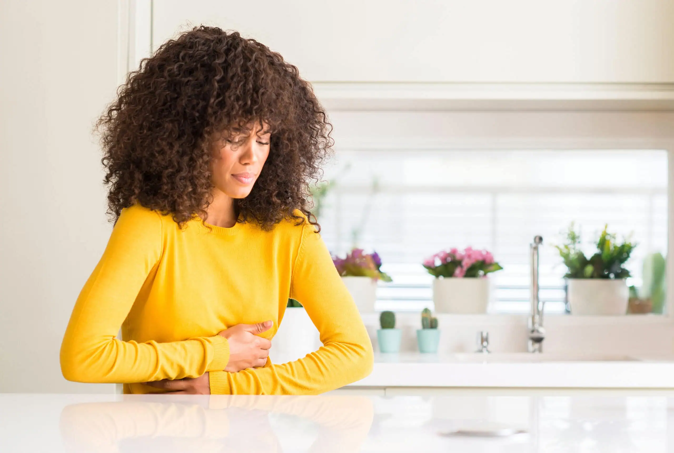 Woman in a yellow sweater holding her stomach and appearing to be in discomfort, standing in a bright kitchen with plants in the background.