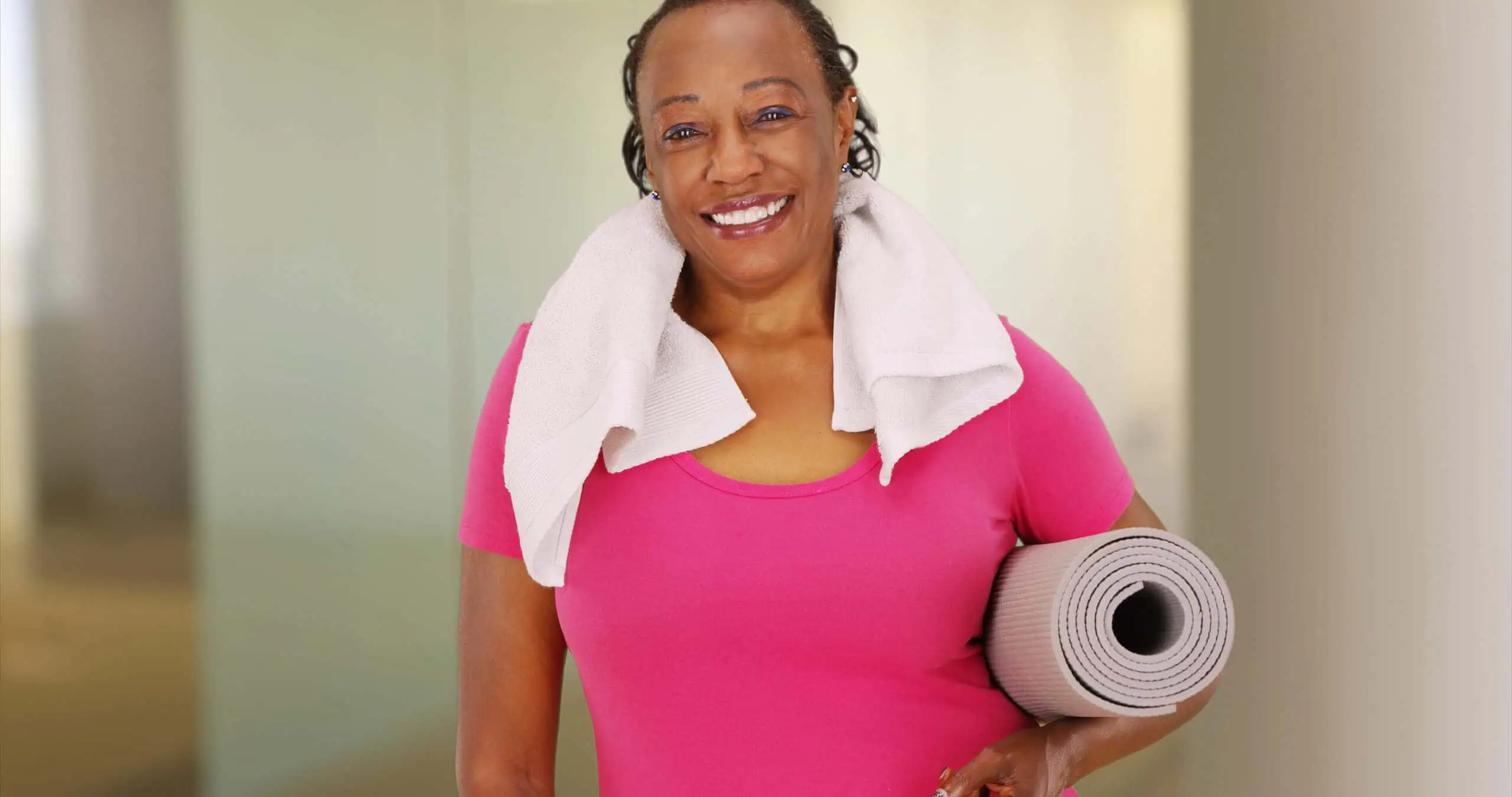 Smiling woman in a pink shirt holds a rolled-up yoga mat and has a towel draped over her shoulders, standing indoors.