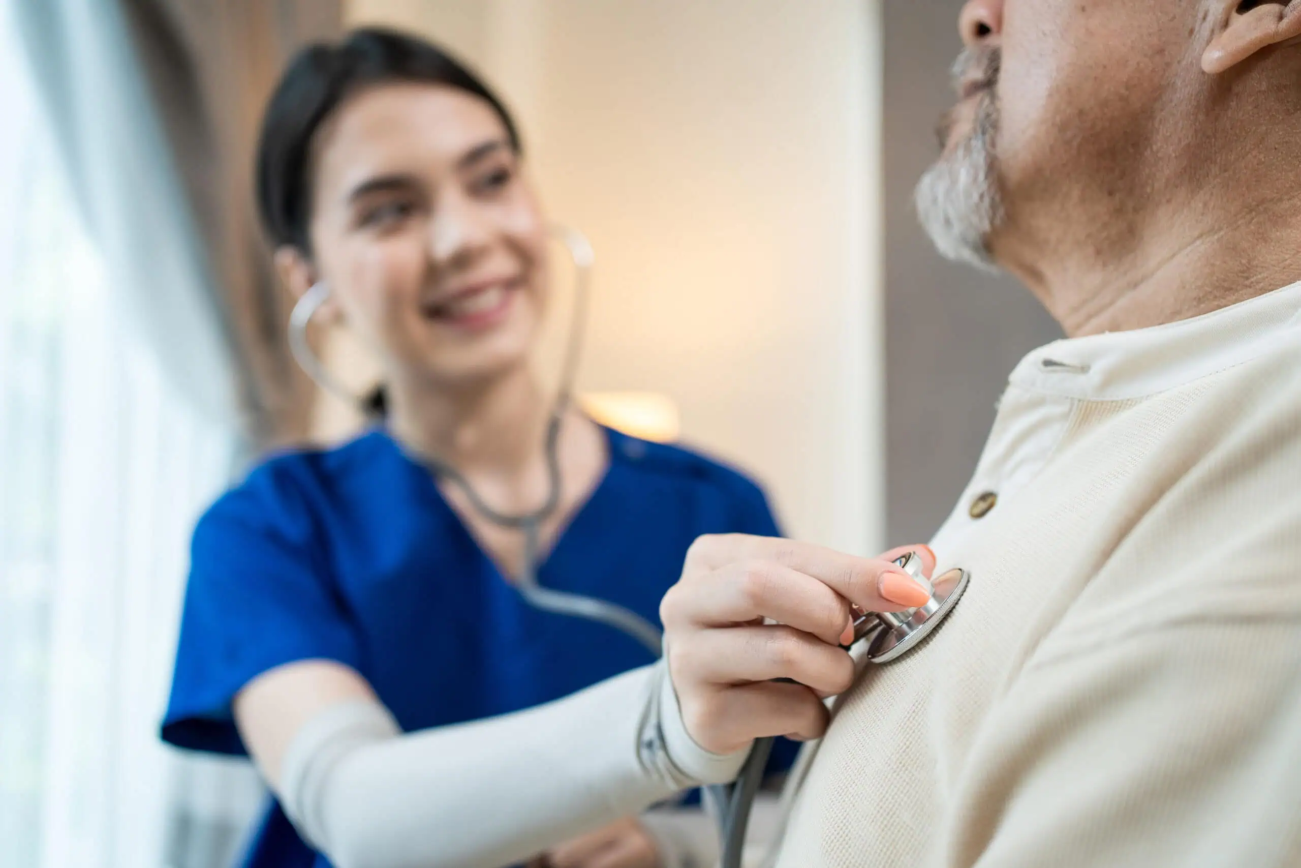 A healthcare worker in blue scrubs uses a stethoscope to listen to an older man's chest during a medical examination.