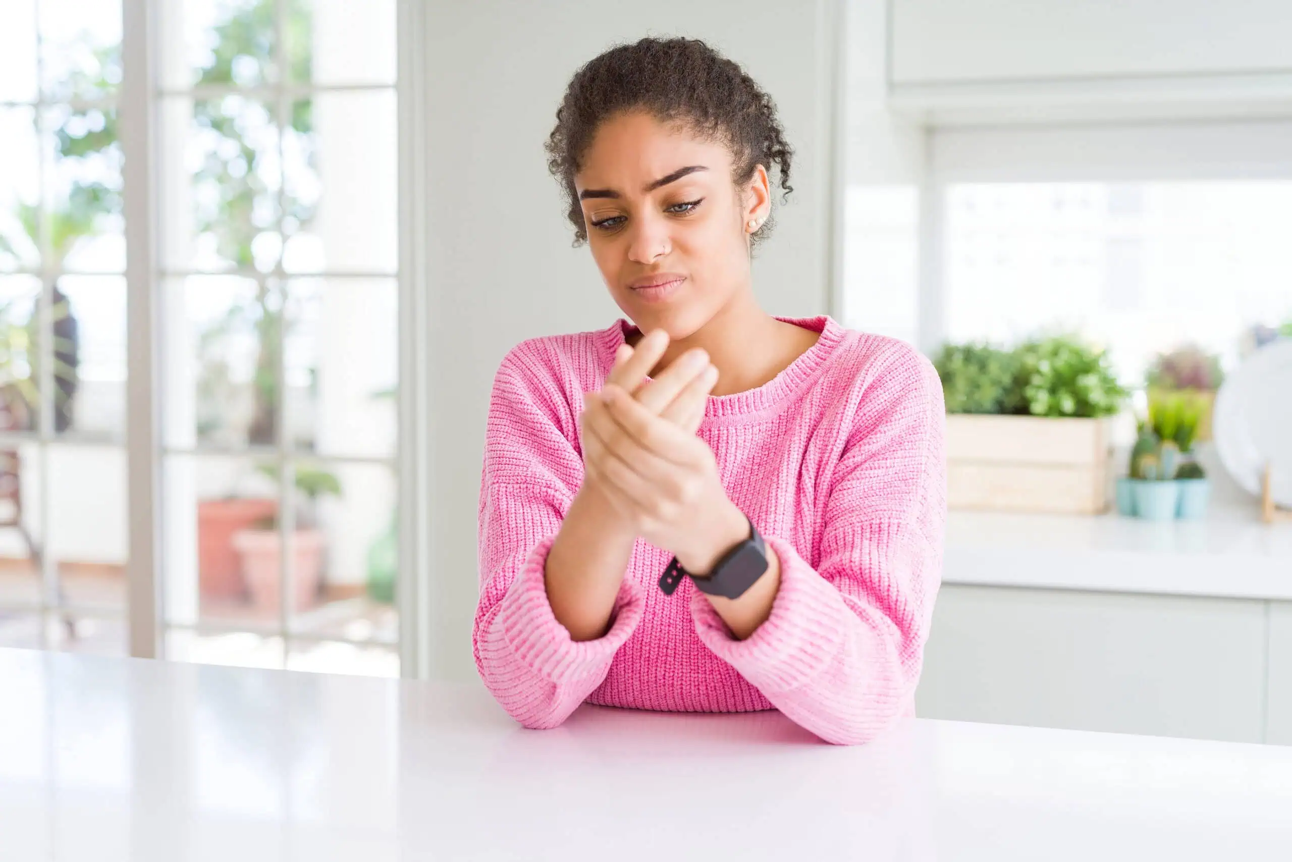 A woman in a pink sweater sits at a table indoors, looking at and holding her painful wrist with a concerned expression.