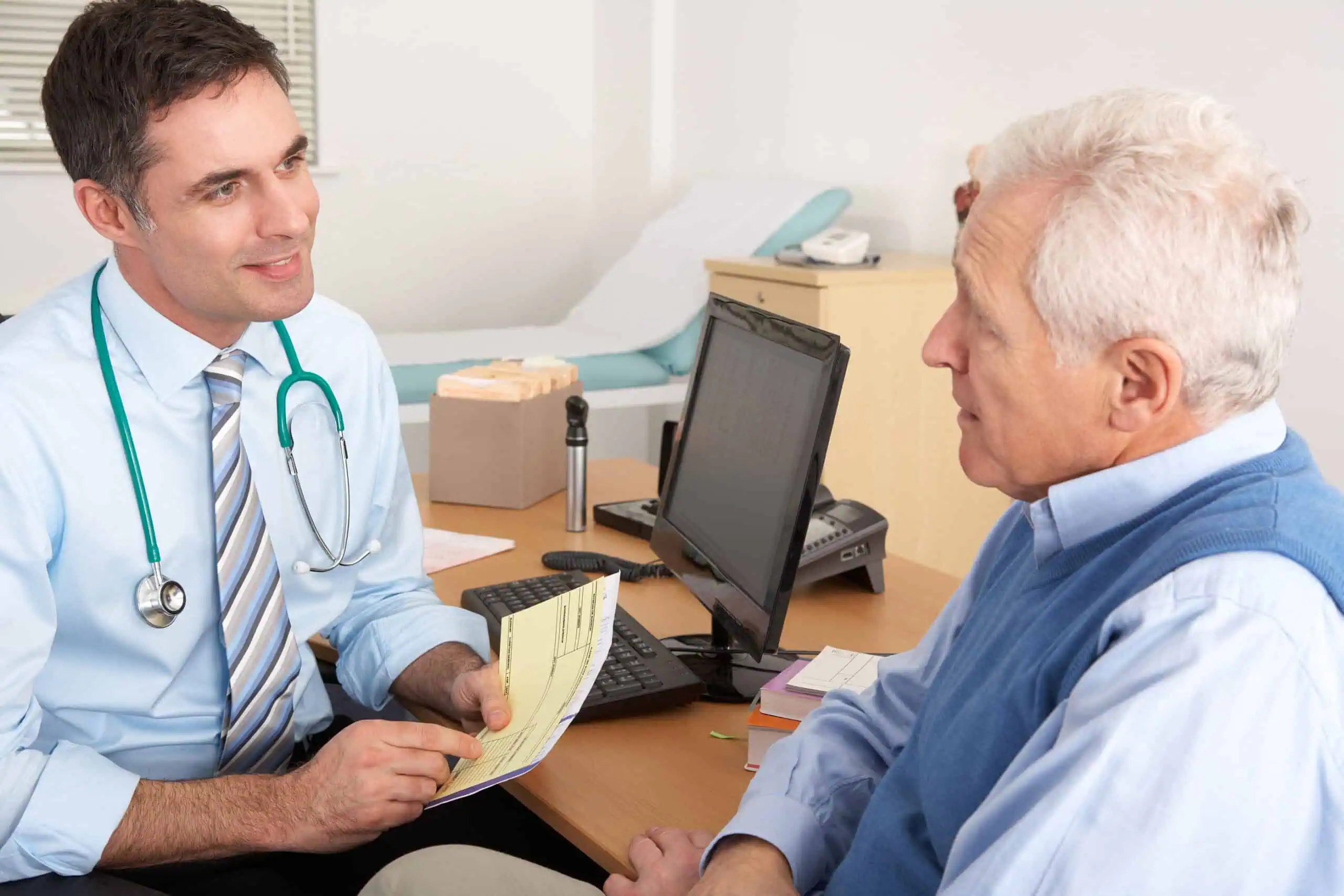 A doctor sits at a desk speaking with an older male patient, reviewing documents together in a medical office setting.