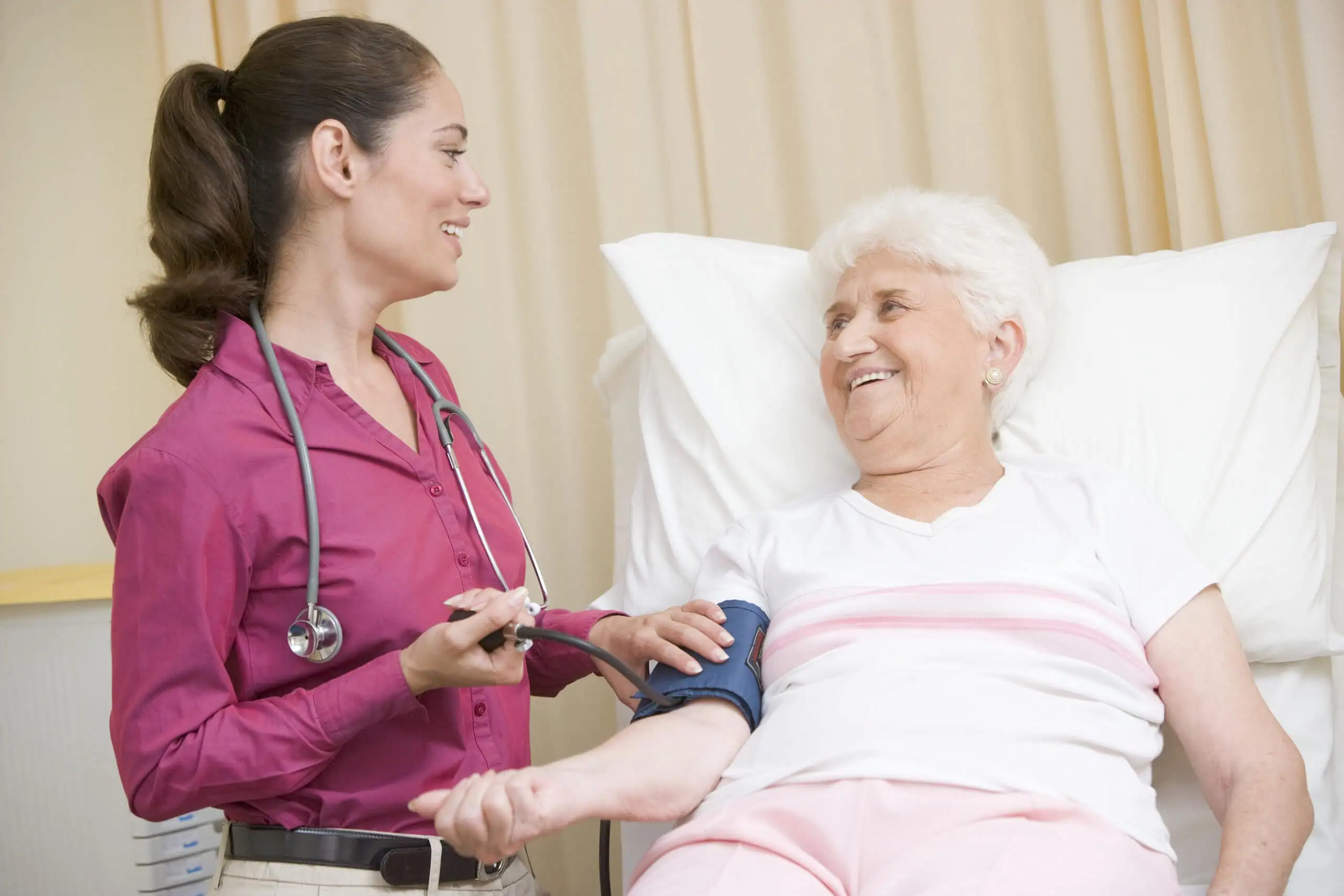 A healthcare worker checks the blood pressure of an elderly woman who is sitting on a hospital bed. Both are smiling and facing each other.