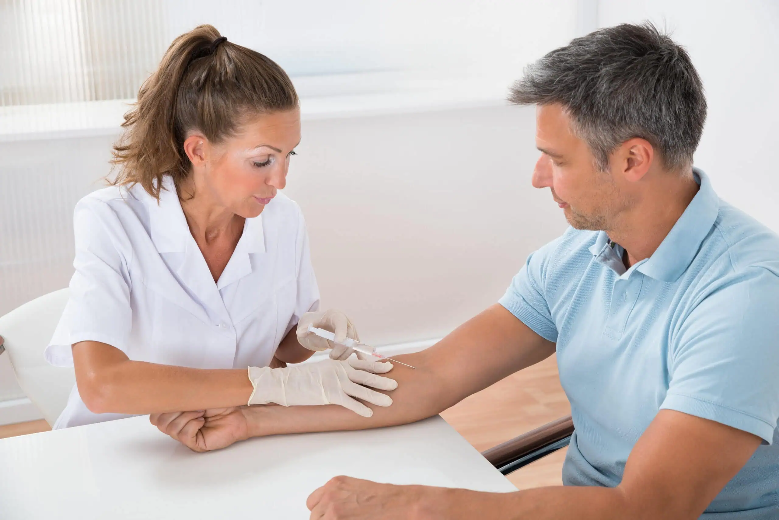 A healthcare professional in white gloves prepares to draw blood from a man's arm using a syringe in a medical office.