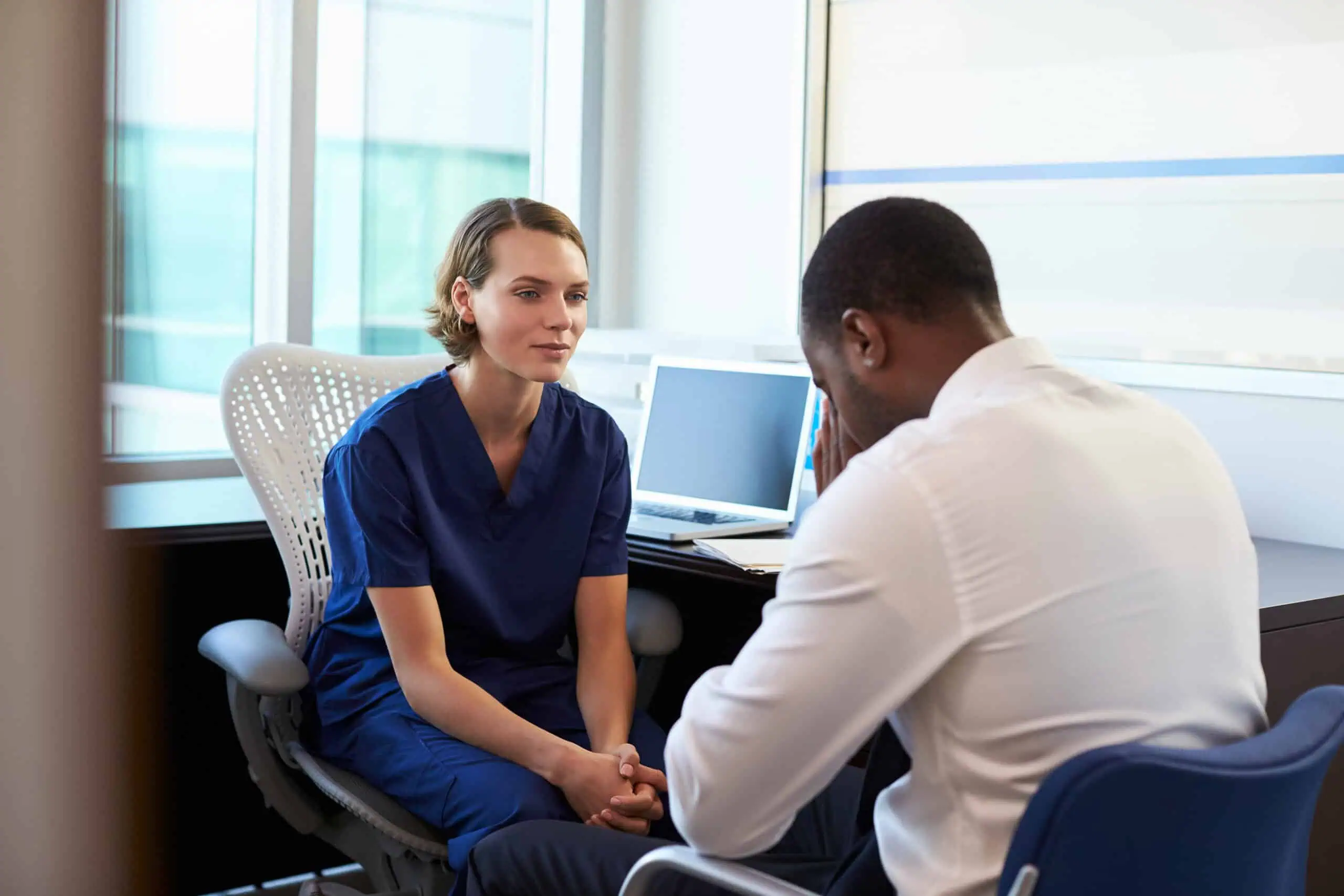 A healthcare professional in scrubs listens to a man in business attire sitting across from her in an office setting.