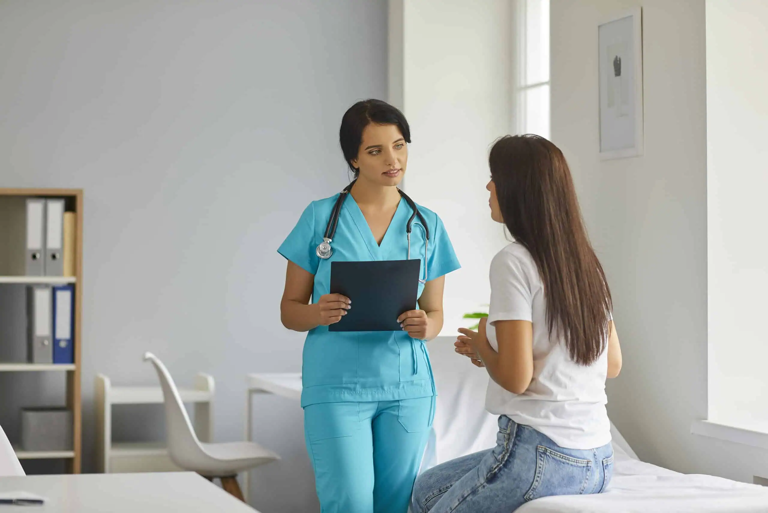 A healthcare professional in blue scrubs holds a clipboard while talking to a patient seated on an exam table in a medical office.