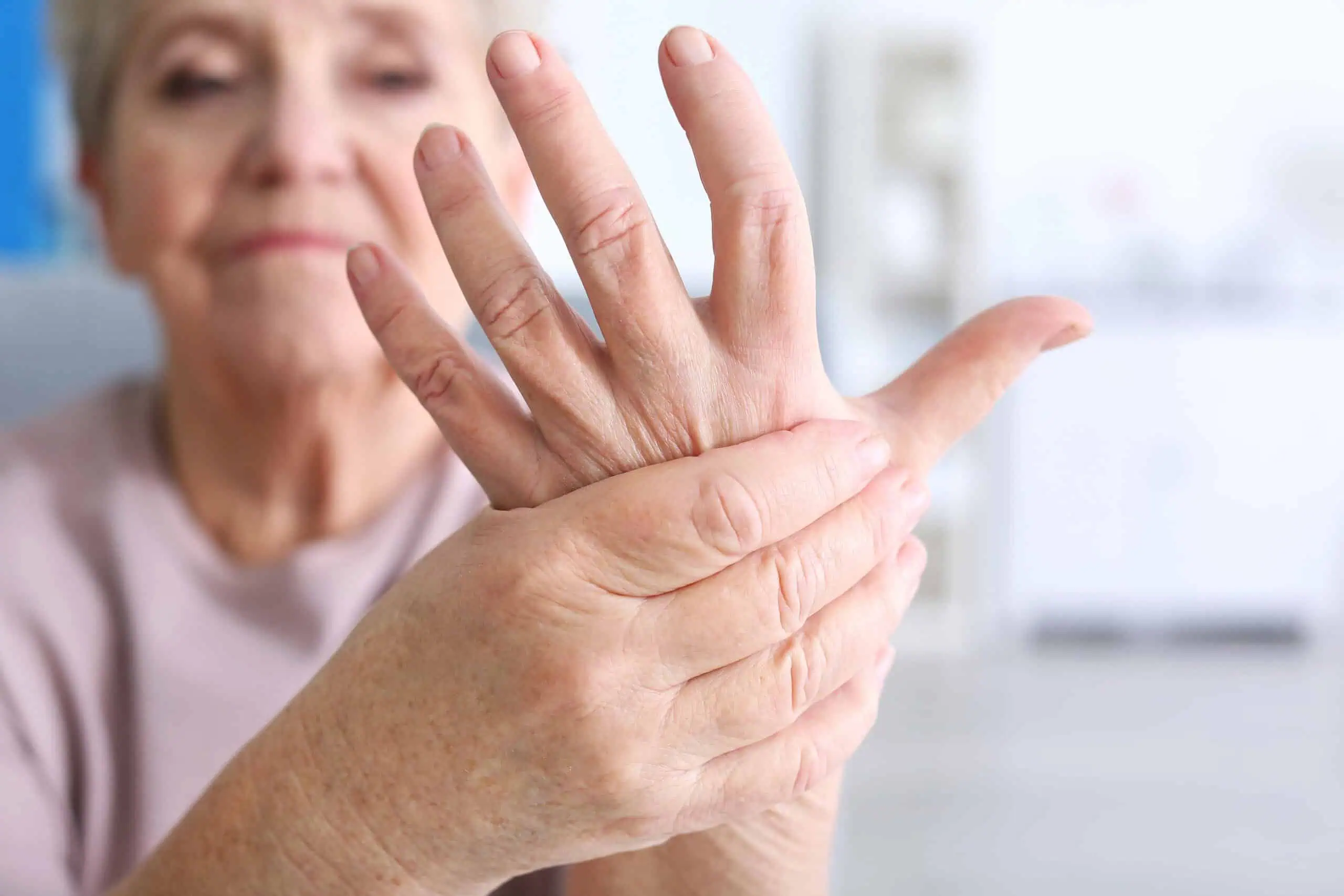 An older woman holds her left hand with her right hand, possibly indicating pain or discomfort in her fingers or joints.