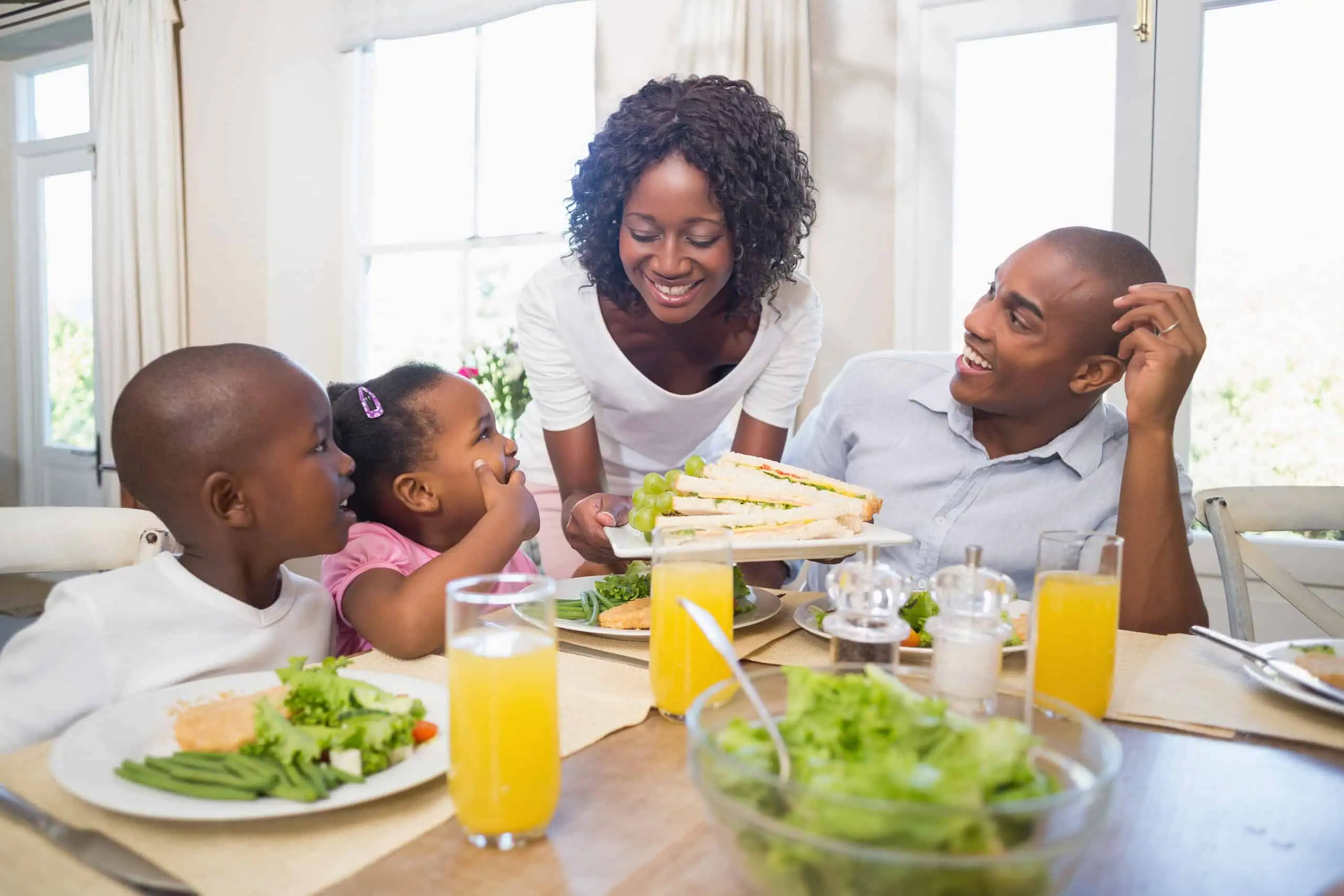 A woman serves sandwiches to a man and two children seated at a dining table set with salad, vegetables, and glasses of orange juice.