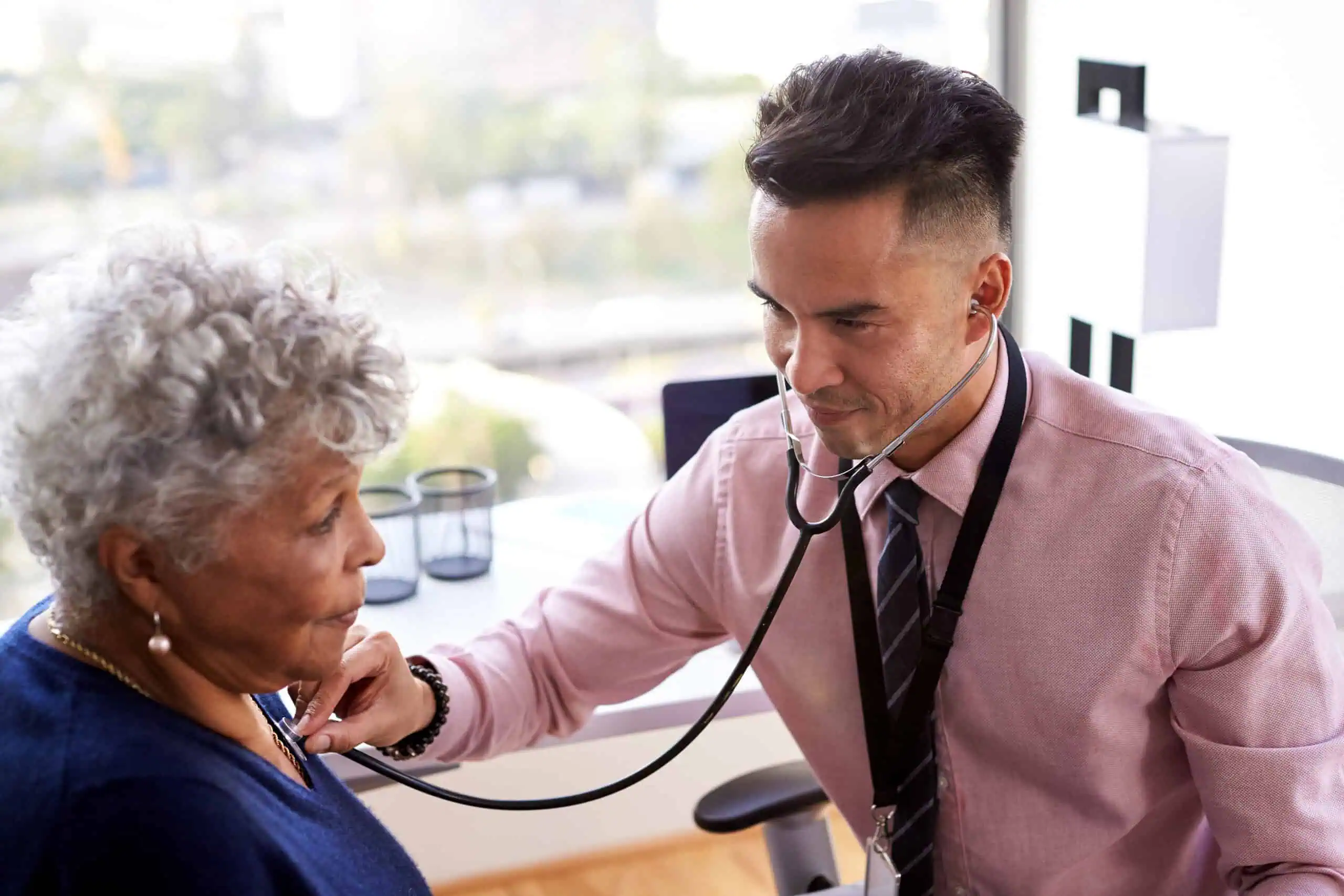 A doctor uses a stethoscope to listen to an older patient's chest during a medical examination in a well-lit office.