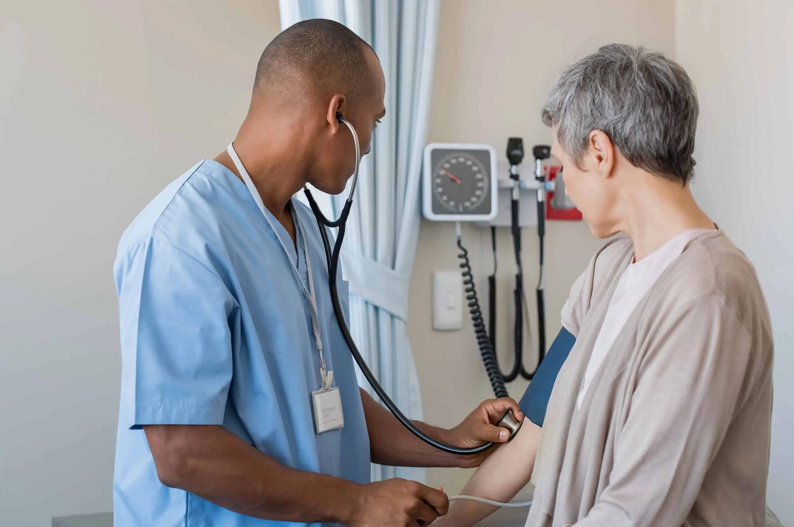 A healthcare professional in scrubs checks a patient’s blood pressure with a stethoscope and sphygmomanometer in a medical exam room.