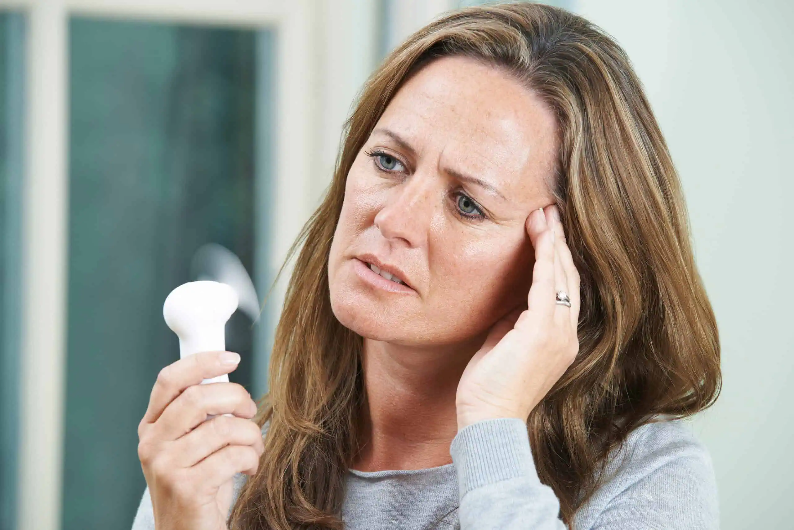 A woman holds a portable fan and touches her forehead, appearing uncomfortable, possibly experiencing heat or a hot flash.