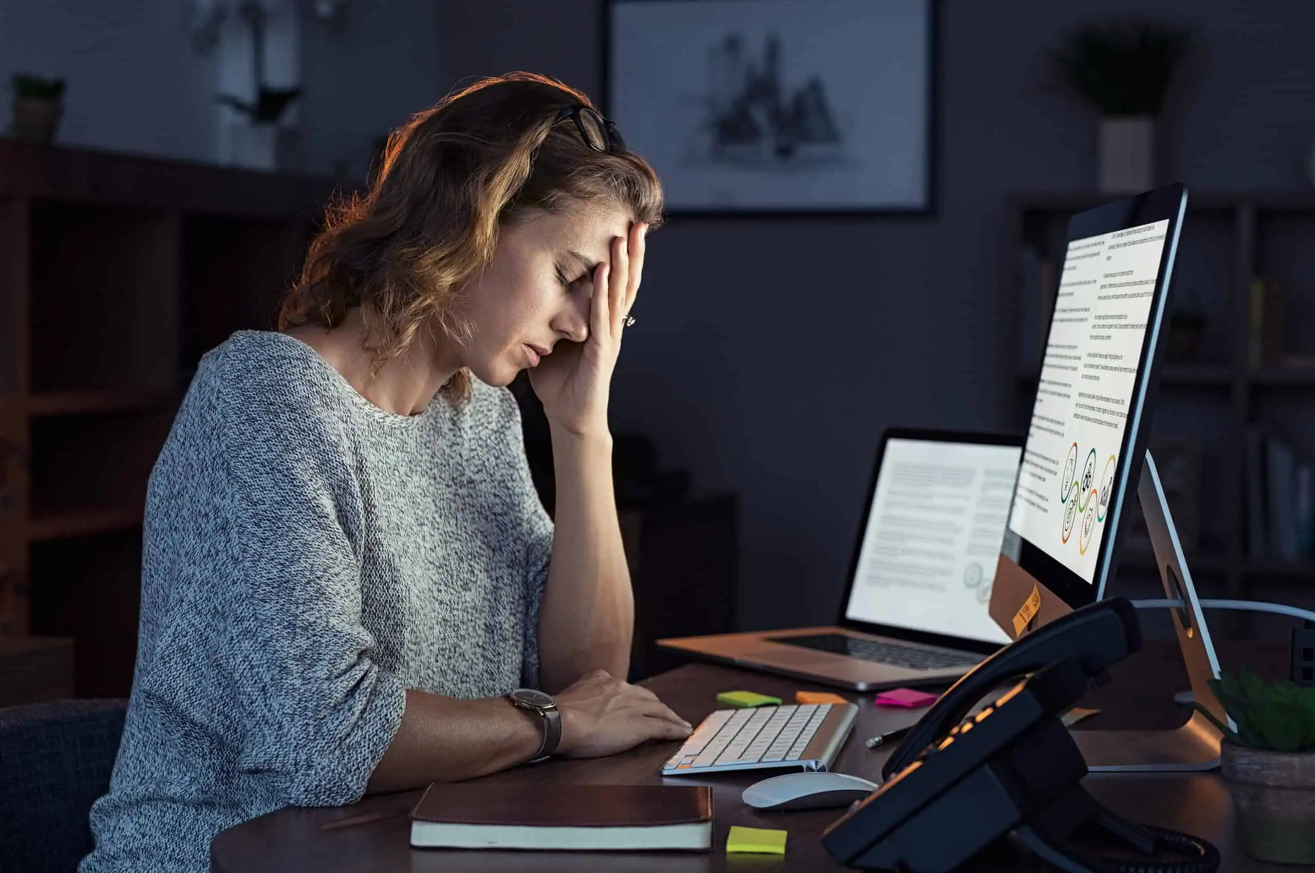 Woman sitting at a desk with two computer monitors, holding her head in her hand and appearing stressed or fatigued while working.