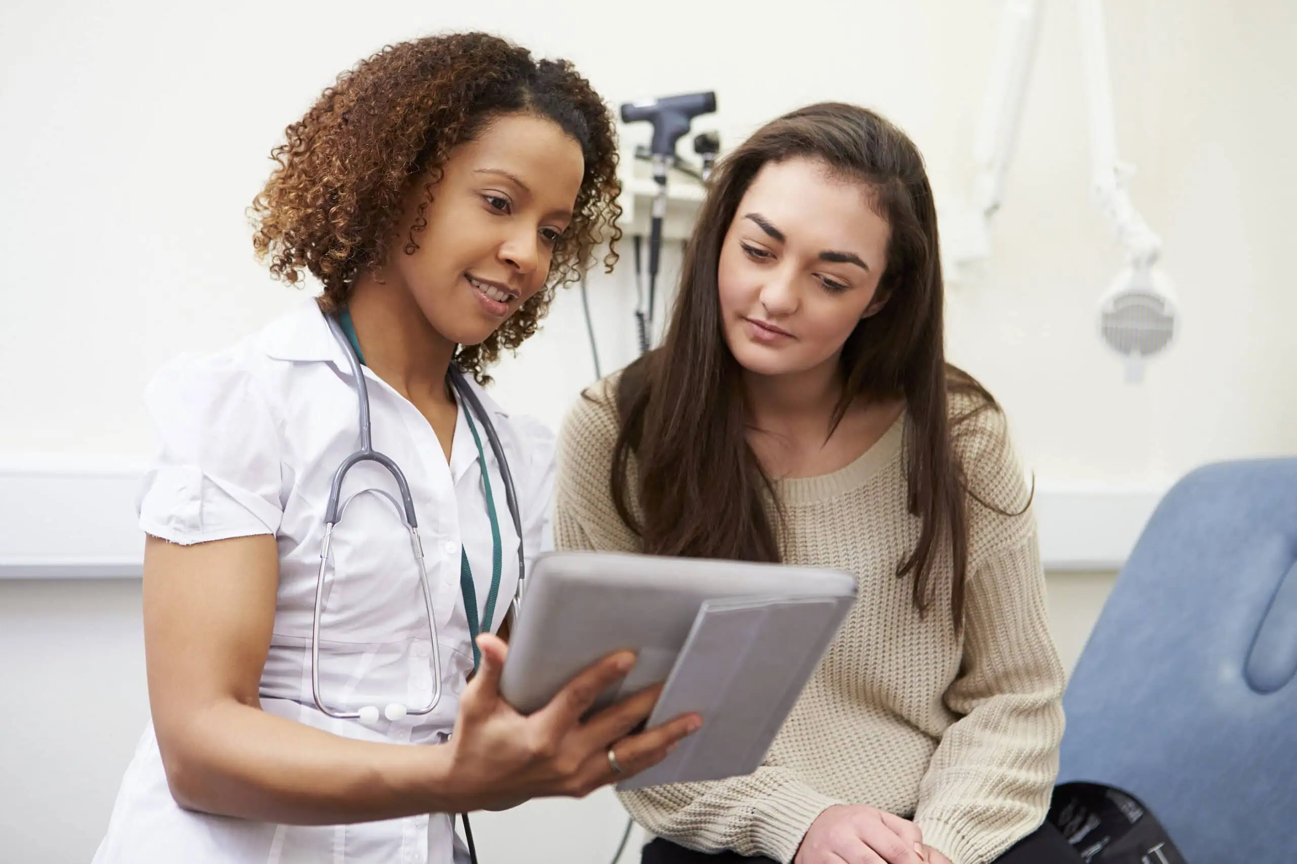 A doctor and a patient sit in a medical office while the doctor shows information on a digital tablet.