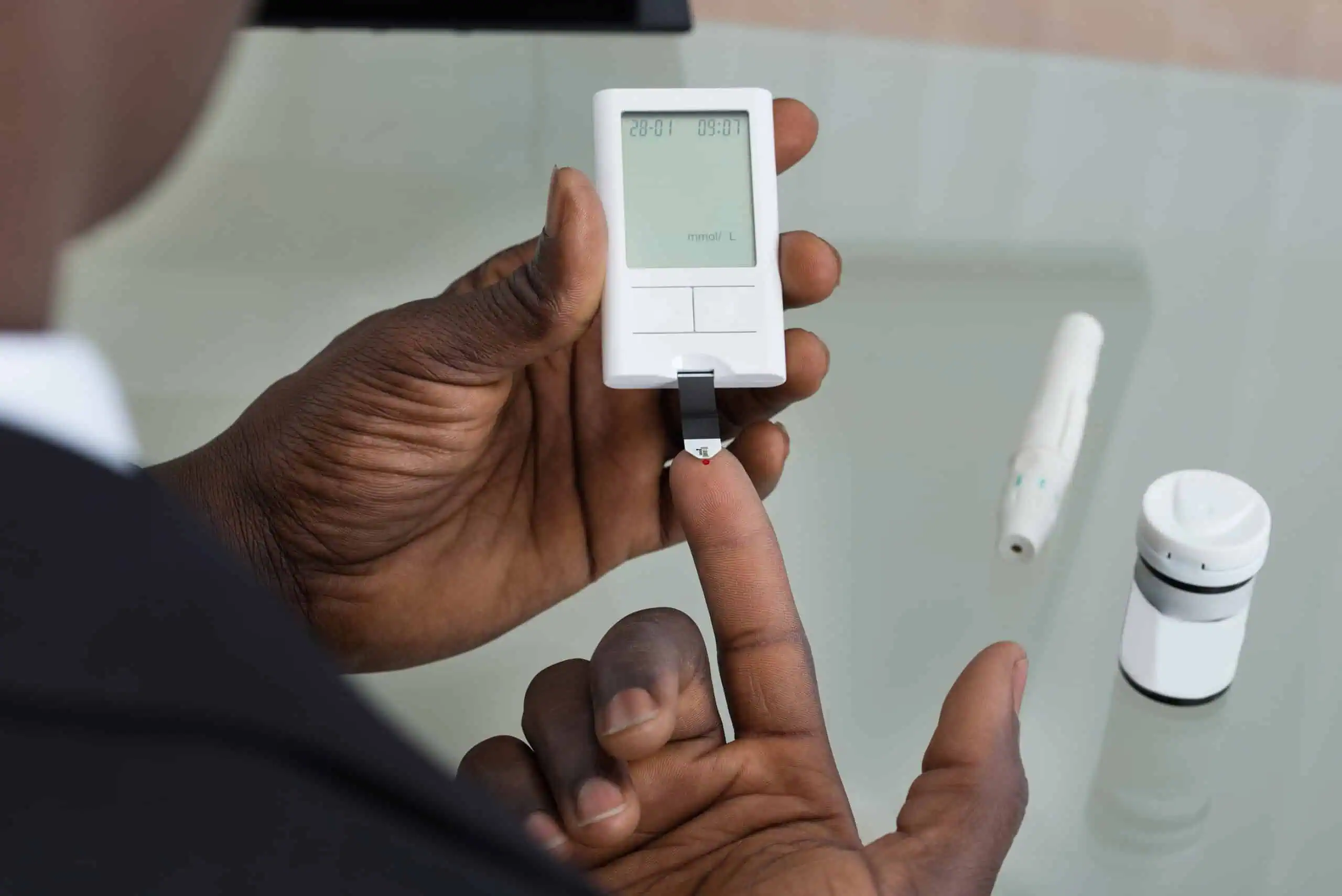 A person uses a glucometer to check their blood sugar level by placing a blood sample on a test strip. A lancing device and test strip container are on the table, demonstrating daily diabetes management as part of Primary Care in Cayman.