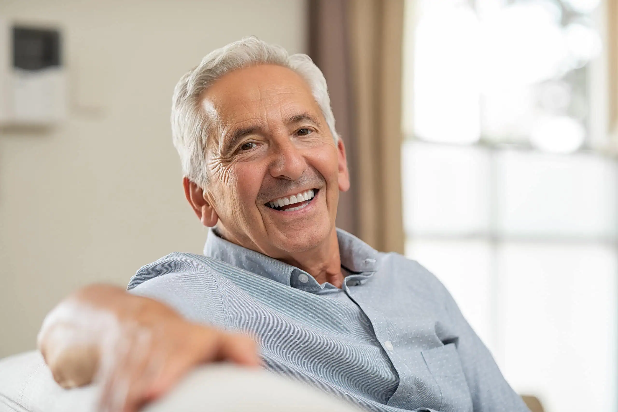 Older man with gray hair and a light blue shirt sits on a couch, smiling, with a window and curtain in the background.
