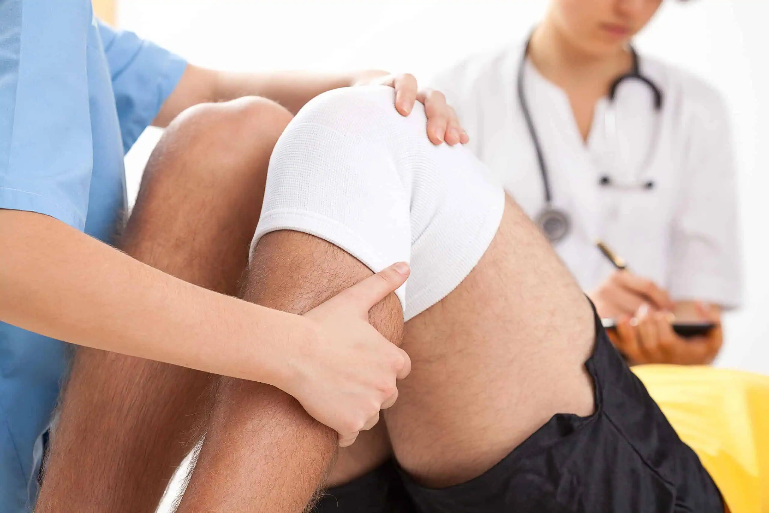 A medical professional examines a patient's knee, which is wrapped in a bandage, while another healthcare worker takes notes in the background.