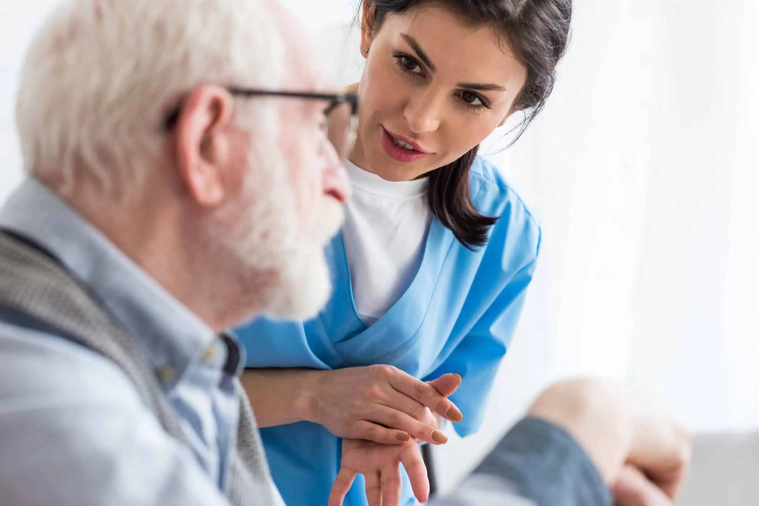 A nurse in blue scrubs attentively listens to an elderly man with white hair and glasses during a conversation.