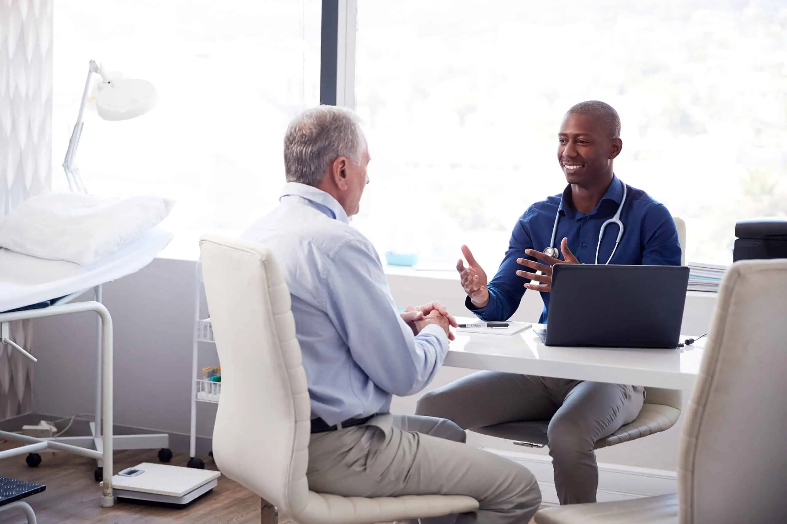 A doctor and a patient sit across from each other at a desk in a medical office, engaged in conversation. A laptop and stethoscope are on the desk.