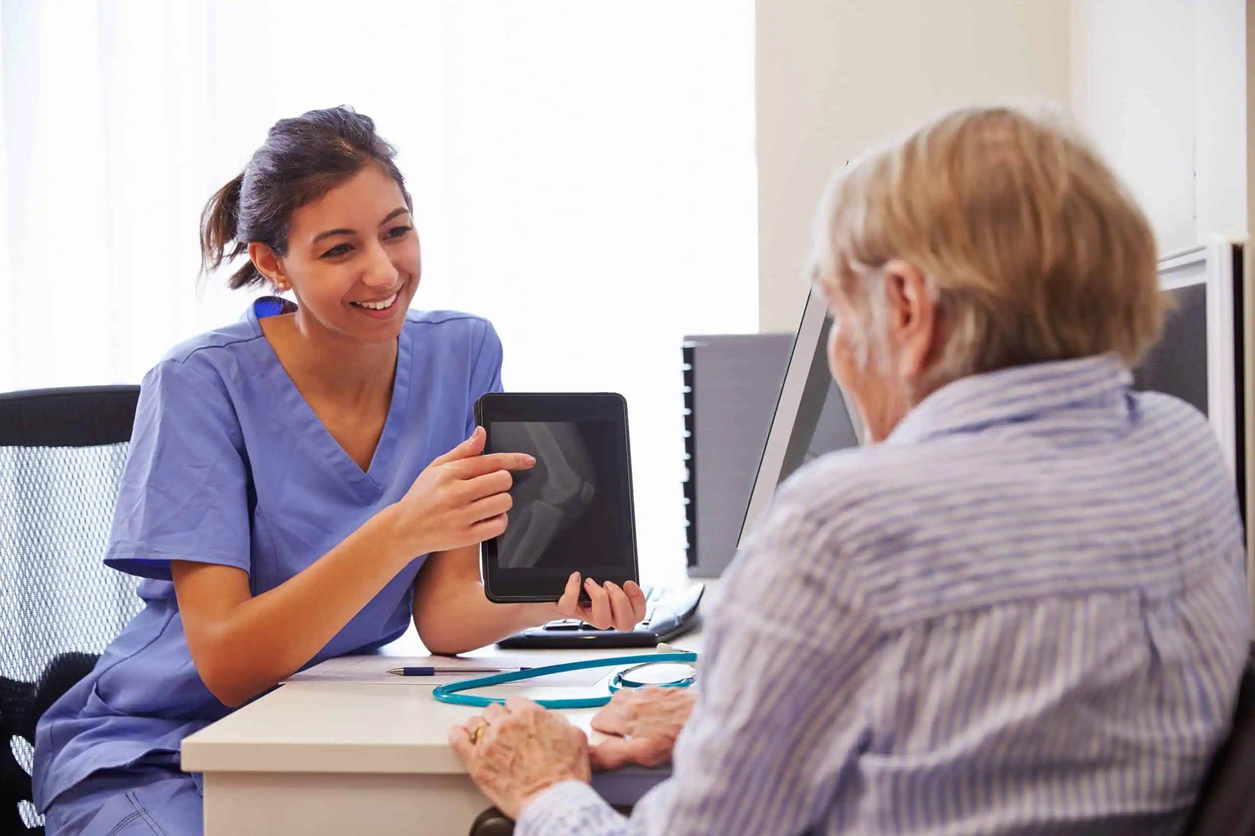 A healthcare professional shows a tablet with a medical image to an older patient during a consultation at a desk.