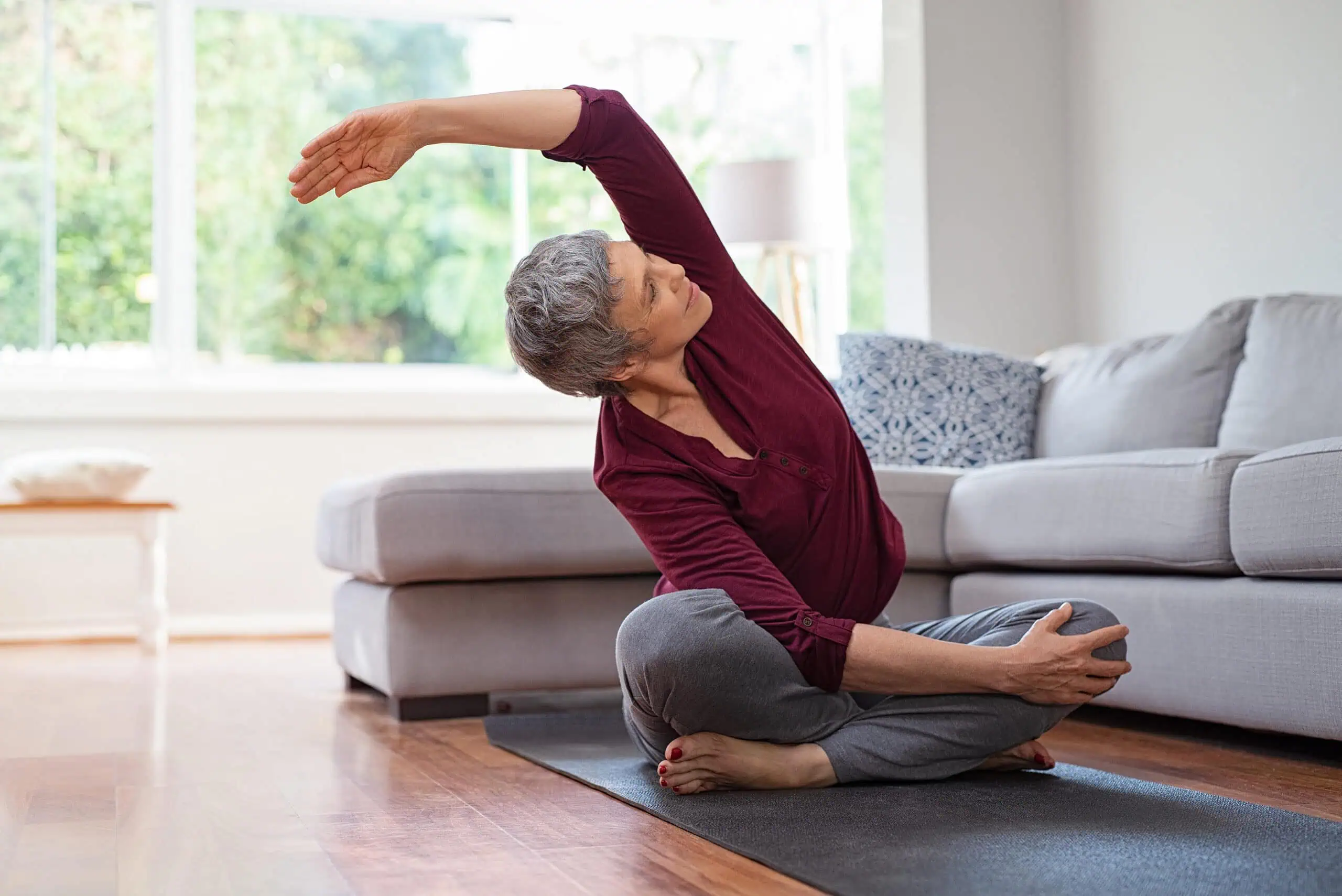 Older adult sitting cross-legged on a yoga mat in a living room, stretching one arm overhead and leaning to the side.
