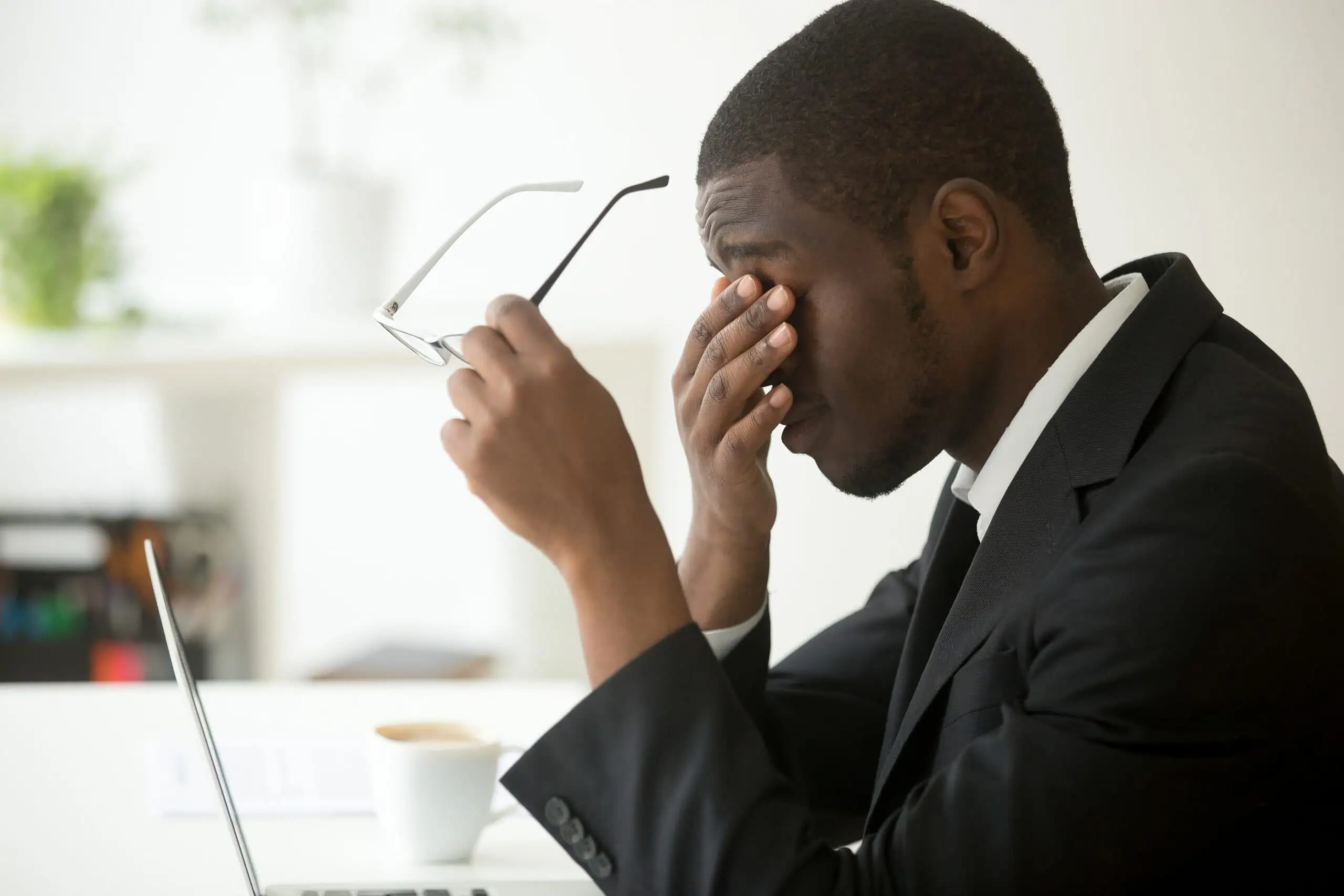 A man in a suit sits at a desk with a laptop, holding his glasses and rubbing his eyes, appearing tired or stressed.