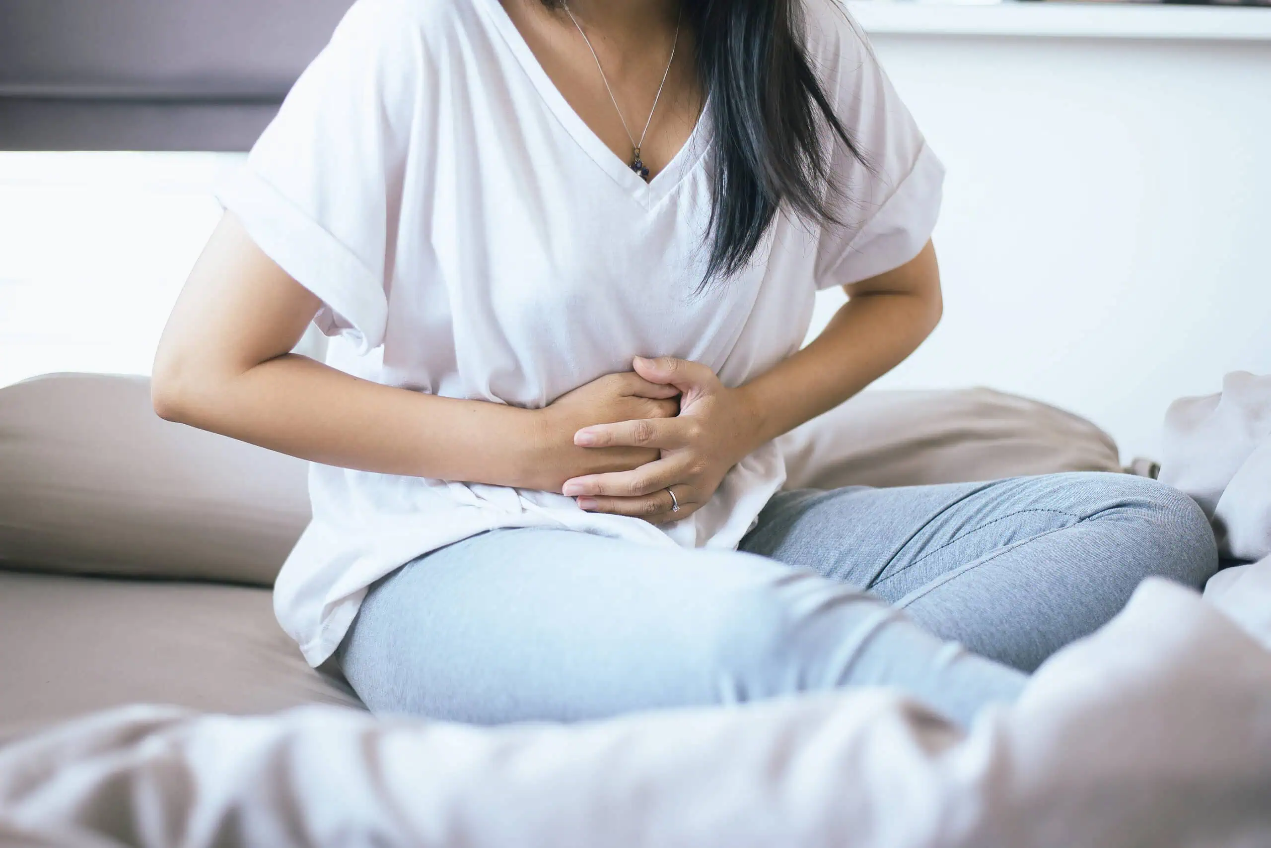 A woman sits on a bed with her hands on her stomach, appearing to experience abdominal pain or discomfort.