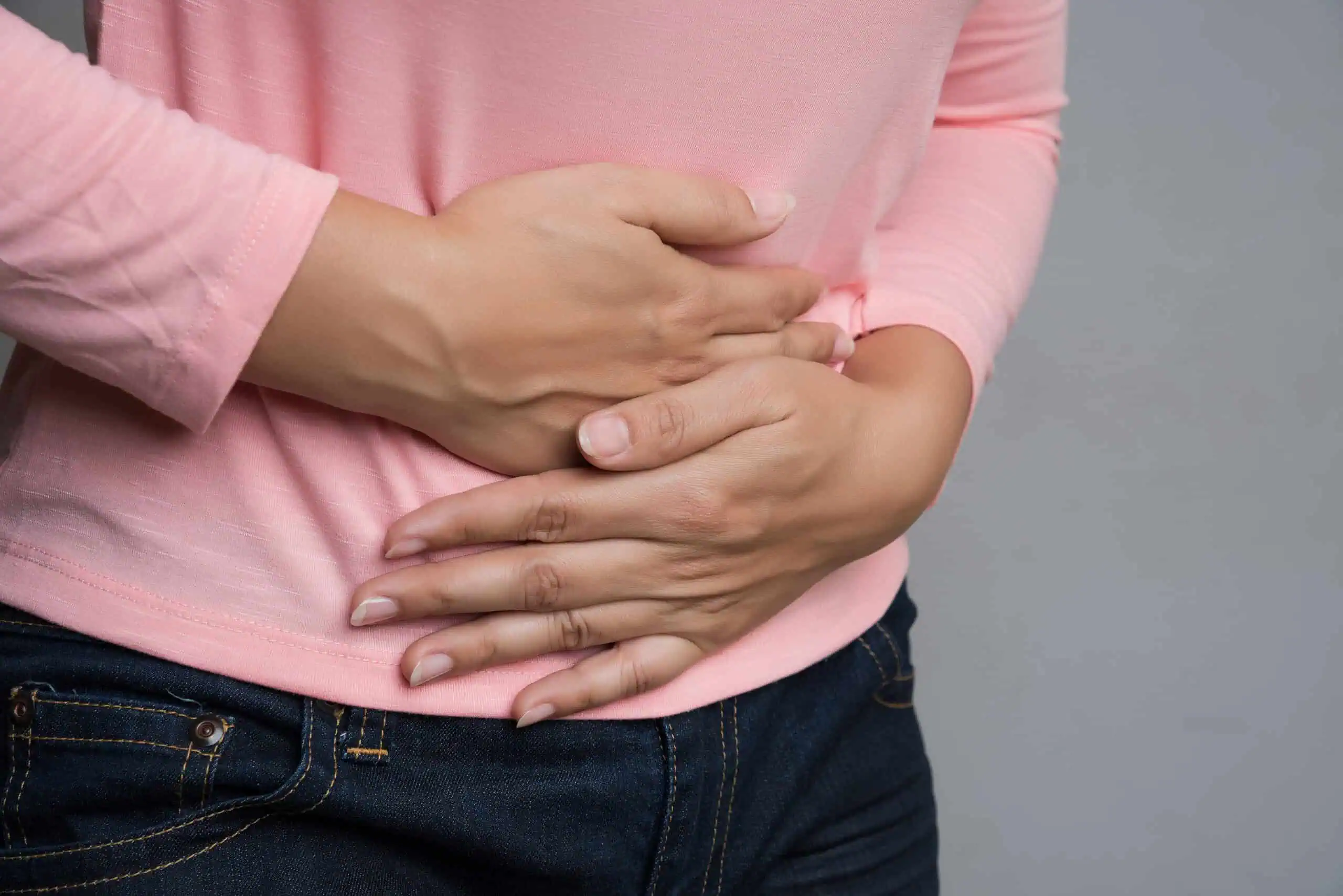 A person wearing a pink shirt and dark jeans holds their stomach with both hands, suggesting discomfort or abdominal pain.