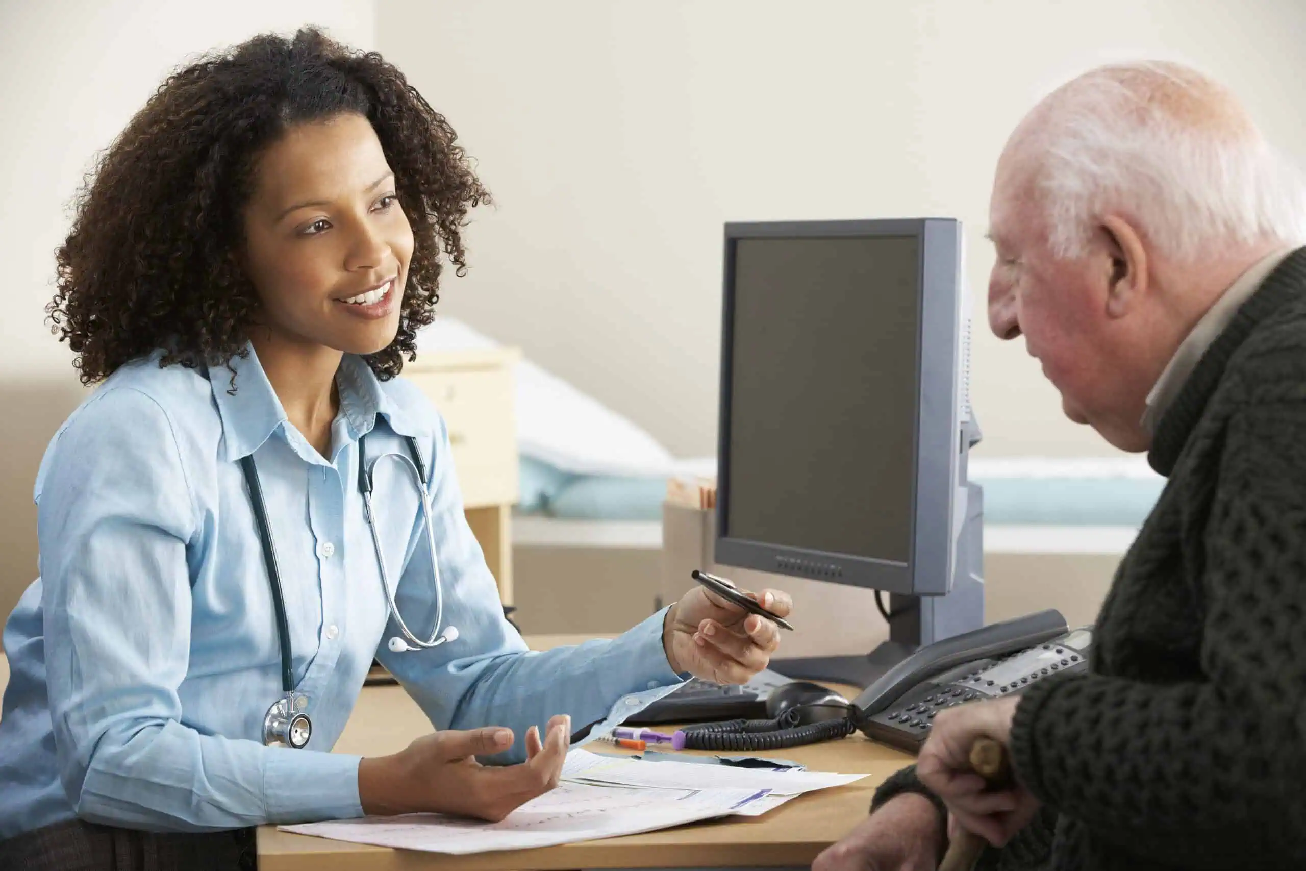 A doctor sits at a desk talking to an elderly male patient, holding a pen and papers, with a computer and phone nearby.